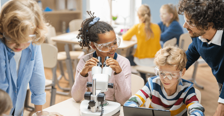 Young black female student with safety goggles on looking in a microscope next to a young white male student with safety goggles on sitting on a laptop. Two teahers stand nearby.