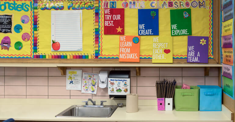 A sink and counter in a STEM classroom. Above the counter, there is a large yellow bulletin board featuring information and motivational phrases. 