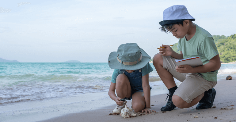 Two children in hats on a beach, crouching over a shell. One child holds a notebook and pencil.