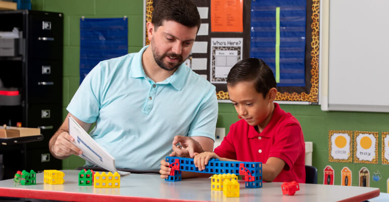 Male teacher in a light blue polo sitting beside a young student in a red polo as they work together on building a structure out of blocks.