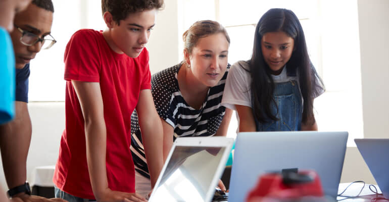 Middle school teacher with a few students leans over a desk, which has open laptops in front of the students