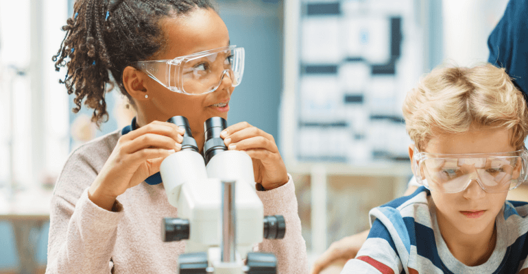 Middle school age Black female student wearing goggle and sitting in front of a microscope. Male student sits nearby.